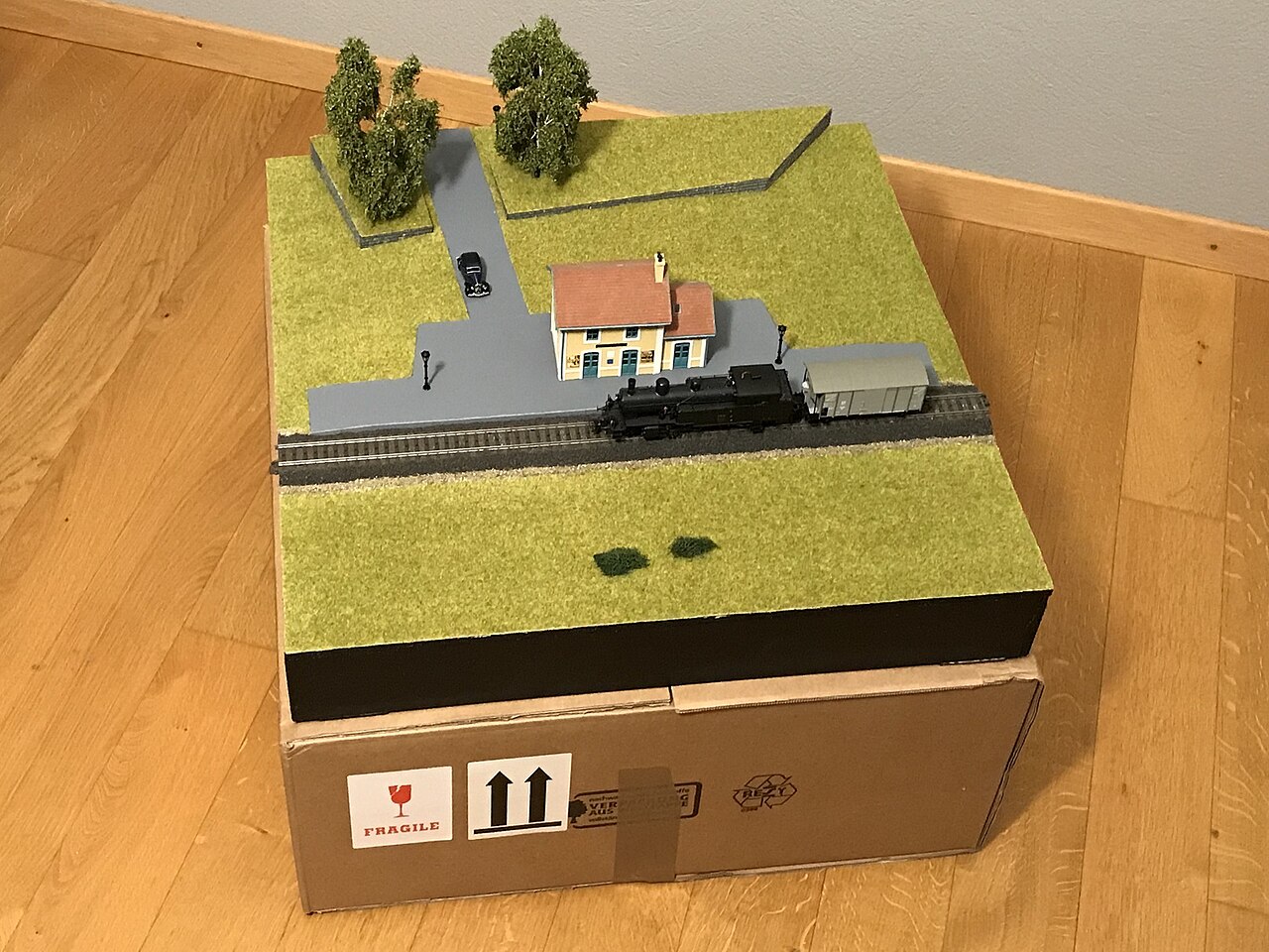 A small H0 module: grass, a road, a small station and trees, with a locomotive and freight car on Märklin C-track, standing on a cardboard box