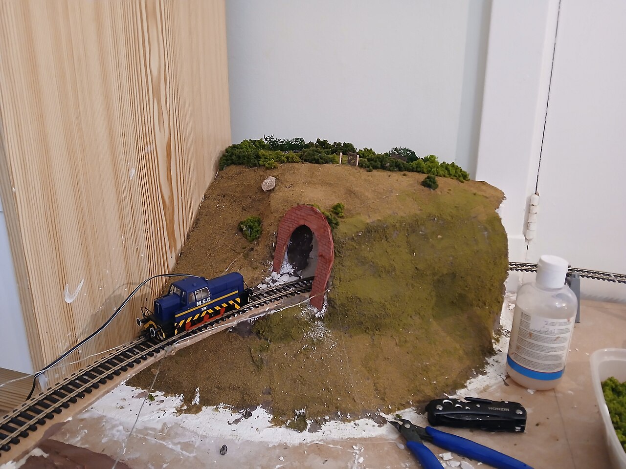 A half-built mountain with a brick tunnel portal, a blue locomotive standing on powered track, with glue bottles and tools beside it