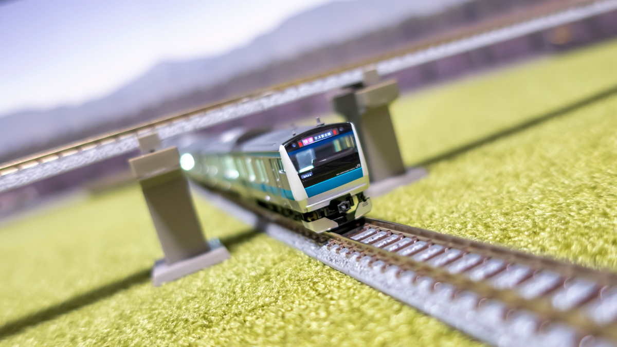 A commuter train passing under a grade-separated crossing on a green flocked board, with an elevated track above and a photo of mountains as the backdrop