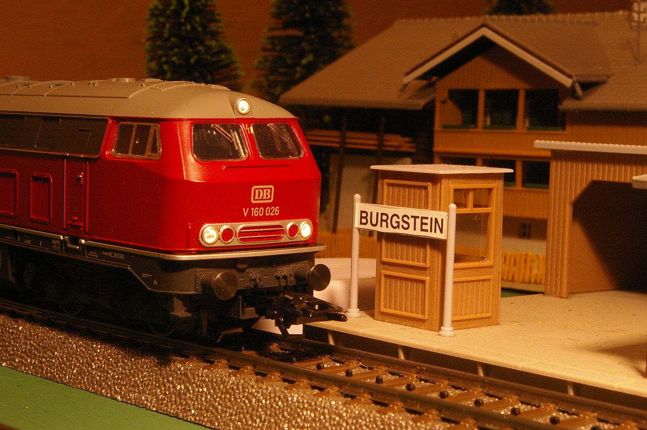 A red V160 diesel locomotive on C-track, with a platform, a station sign and a small building beside it