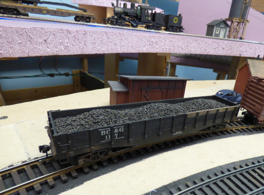 A layout with a foam board as its top: a steam locomotive and freight car on the upper level, a coal gondola and small shed in front, wooden supports and wiring visible underneath
