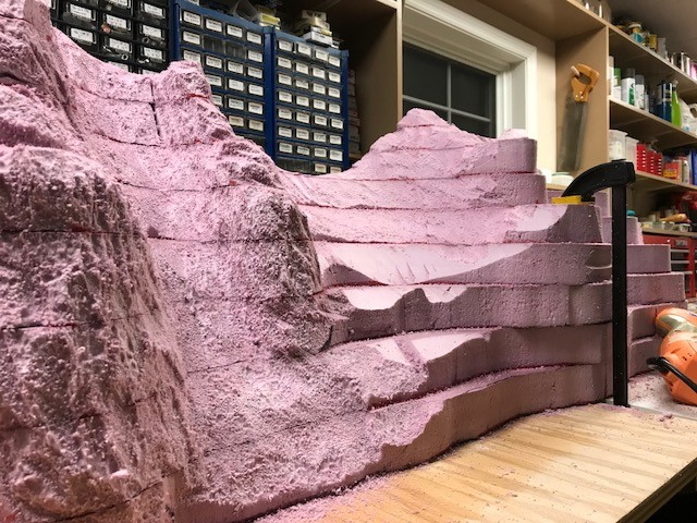 Pink foam board layers stacked and cut into the shape of a mountain, with a clamp beside it and a plywood bench top
