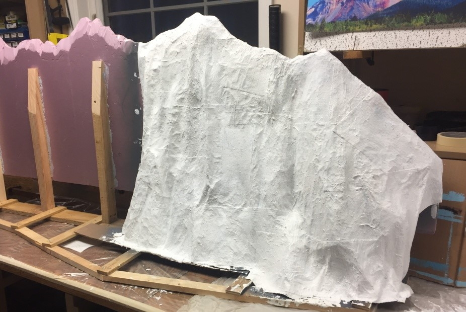A wooden frame covered by a single piece of plaster gauze in the shape of a mountain, with pink foam board visible beside it