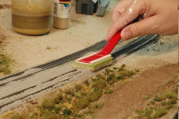 A hand with a red brush tidying grey ballast along both sides of the track, with a glue bottle and ground cover nearby