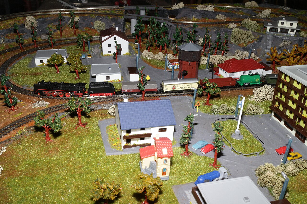 A Z-scale layout: a steam train on a loop, with houses, trees, roads and cars around it, the whole board very compact