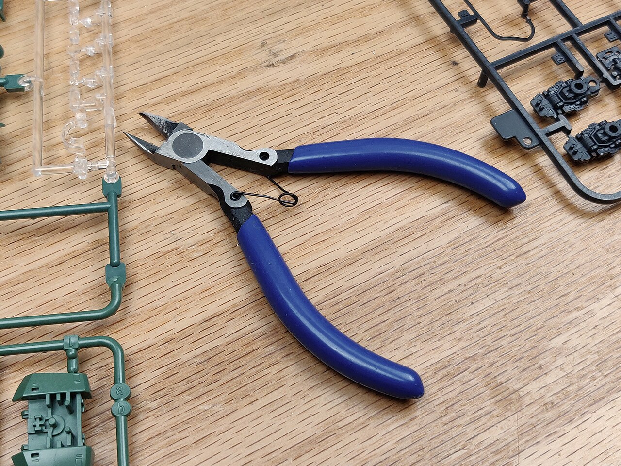 A pair of hobby nippers with a blue handle on a wooden table, surrounded by green, clear and black plastic sprues